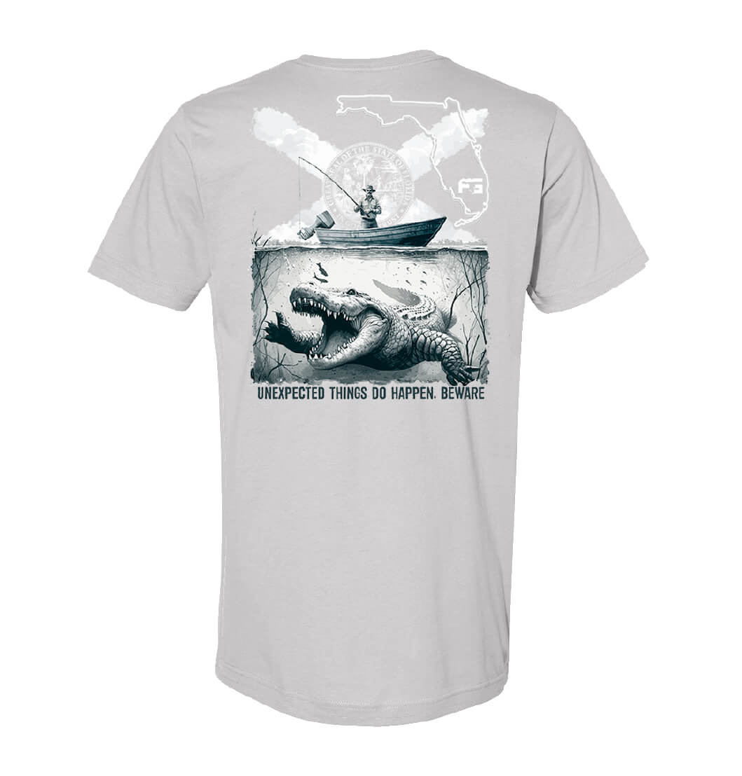 Gray t-shirt with a graphic of an alligator and a boat with text 'UNEXPECTED THINGS DO HAPPEN. BEWARE'.